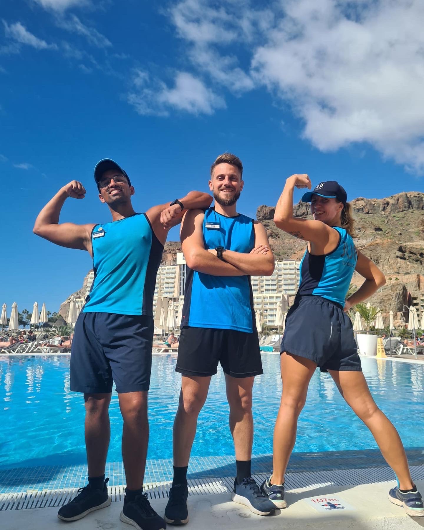 Three fitness instructors stood flexing n front of a pool with blue skies behind.