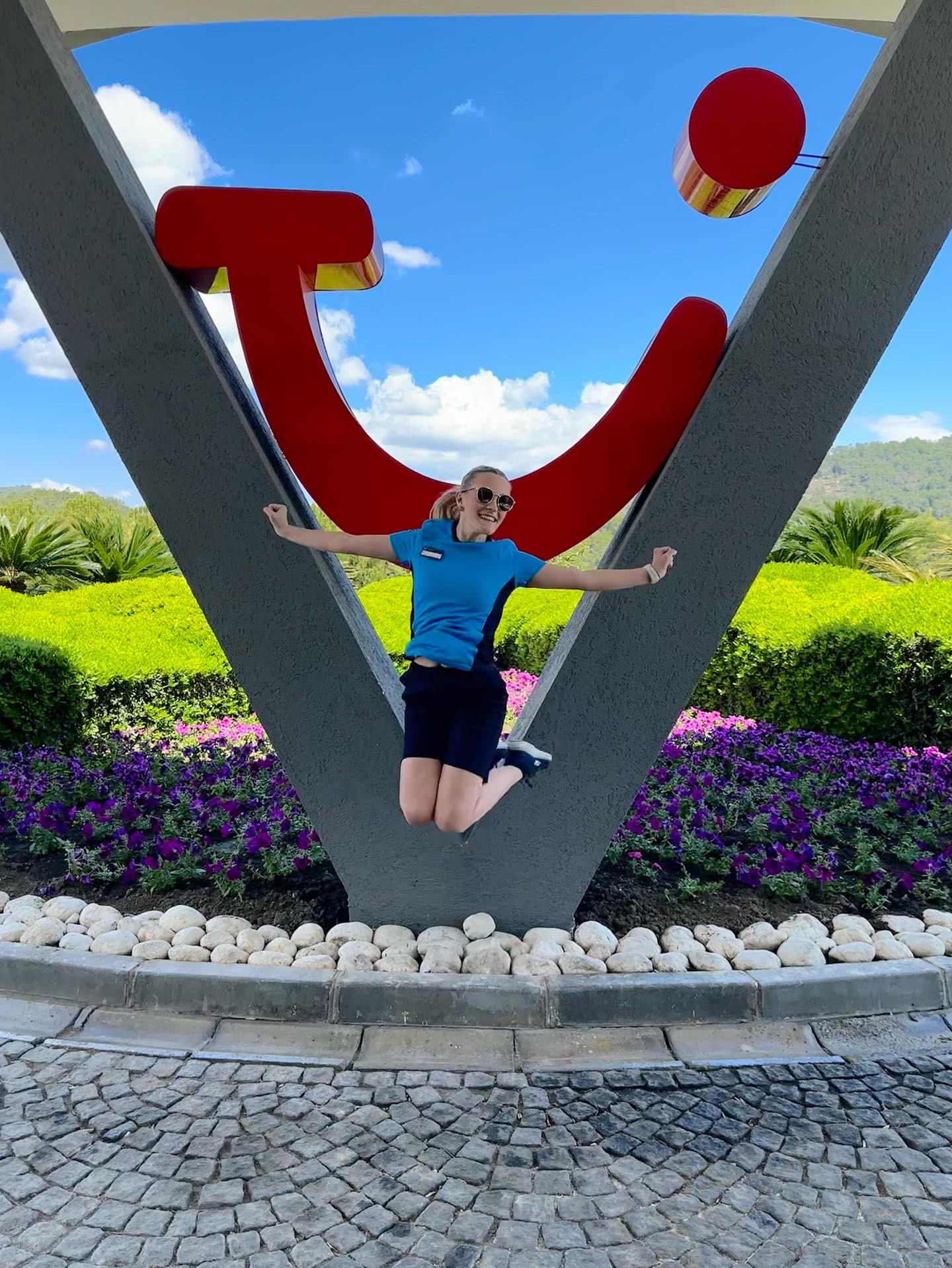 An entertainer is jumping in the air in front of the TUI logo. There is a blue sky in the background and the person is wearing uniform.